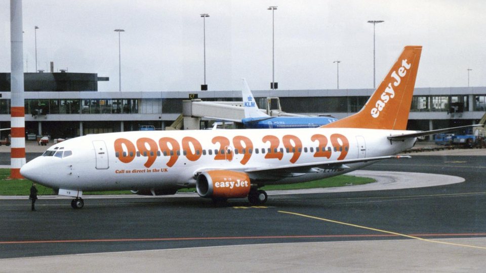 An early-livery easyJet Boeing 737, phone number painted on the fuselage, parked on the tarmac at Amsterdam Schiphol